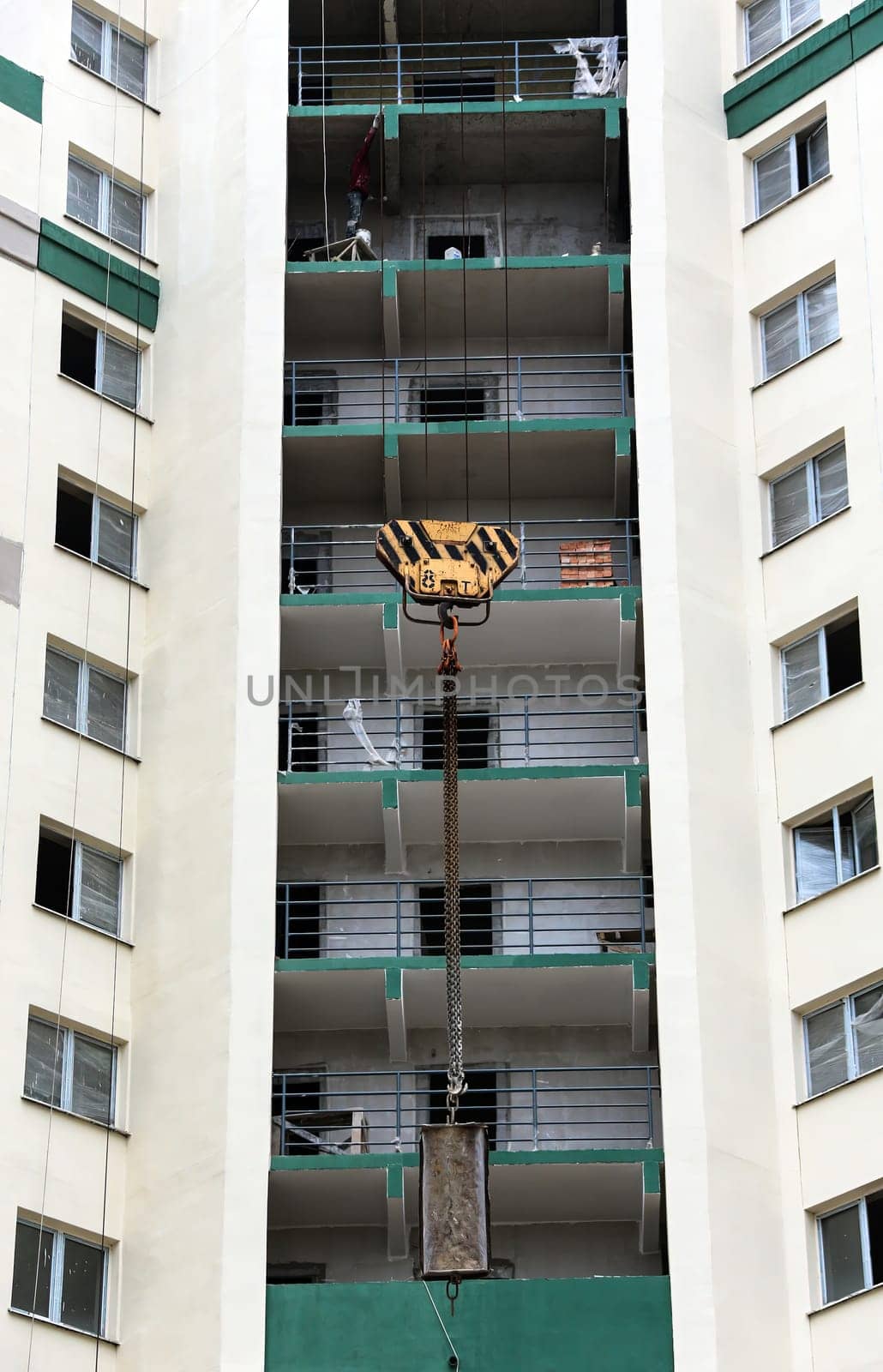 In an urban landscape, a construction crane looms over a high-rise apartment building, diligently lifting heavy materials toward the upper floors. Men can be seen working on the open balconies, focused on their tasks as they prepare for the ongoing construction. The building's exterior, marked by green accents and numerous balconies, reflects the bustling activity of city life.