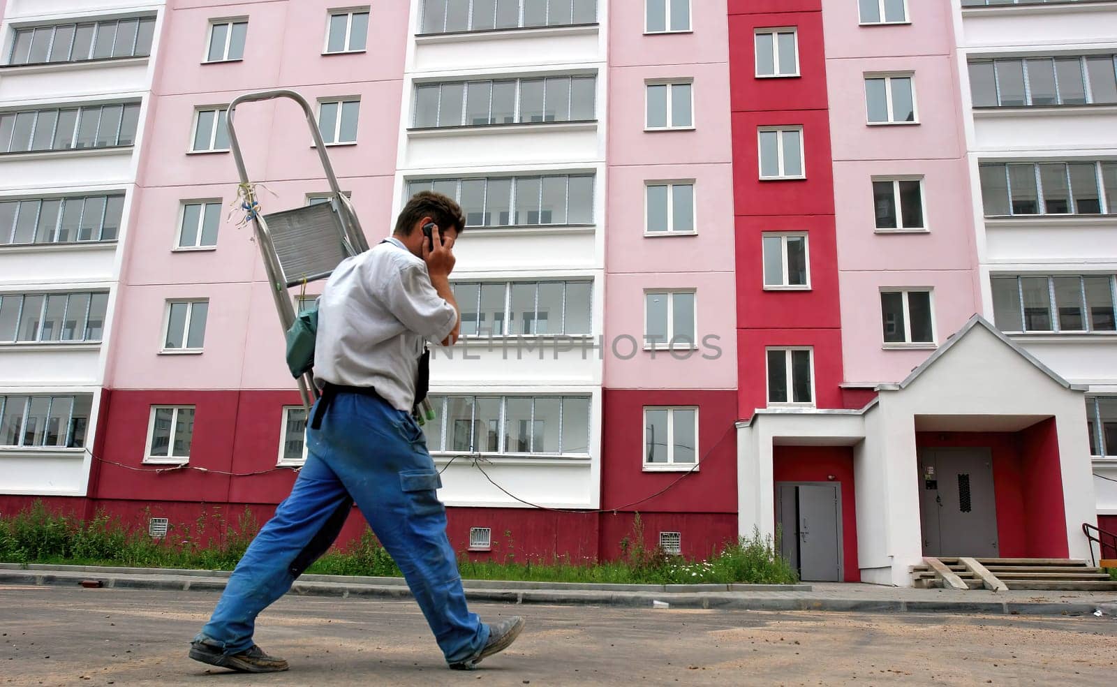A worker with a ladder on his back talks on his phone while walking alongside a newly painted residential building, showcasing vibrant colors and windows.