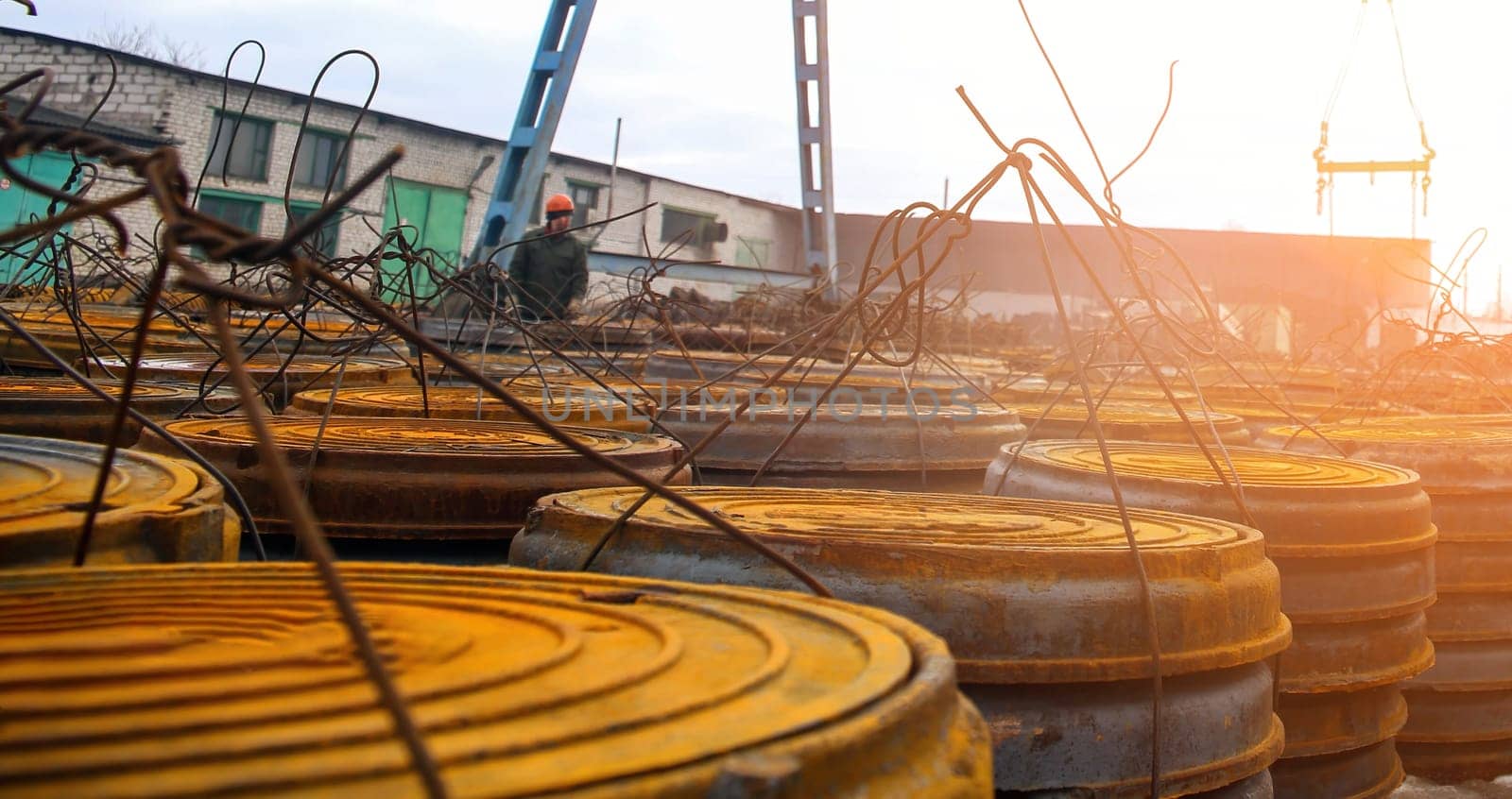 Workers prepare for the next day at a construction site, organizing stacked metal drums surrounded by scaffolding as the sun sets, casting a warm glow.