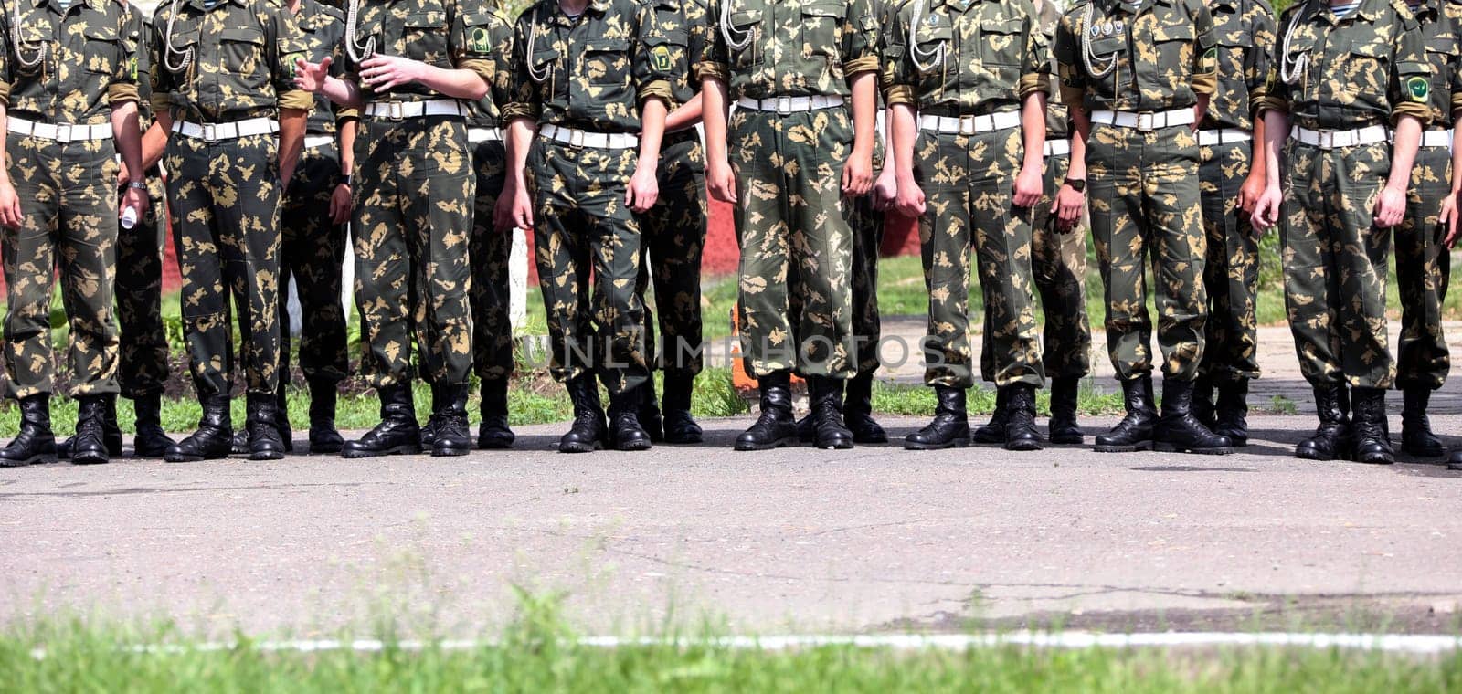 Groups of soldiers in camouflage uniforms stand at attention on a training ground under clear skies during summer.