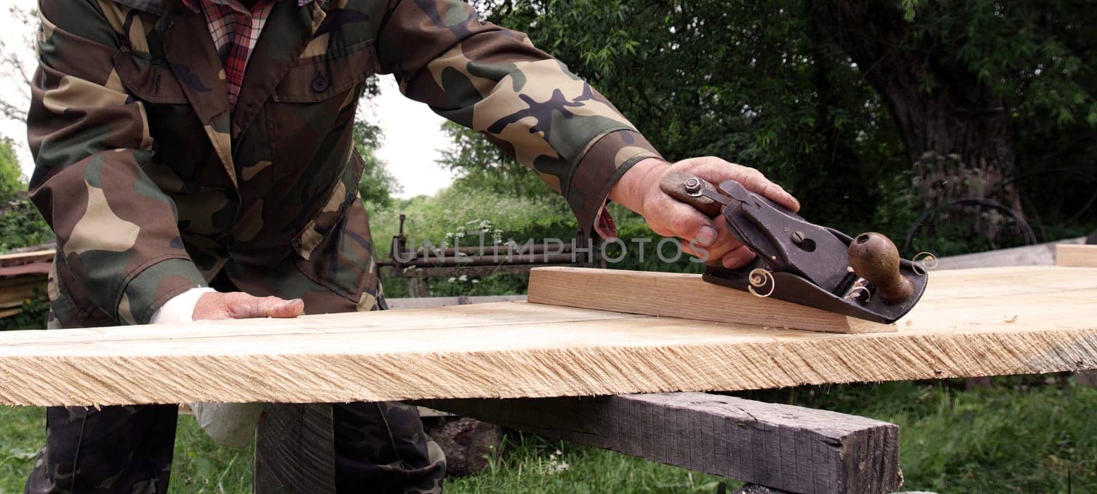 Craftsman works meticulously on a wooden plank using a hand planer in a peaceful outdoor workshop during daylight.