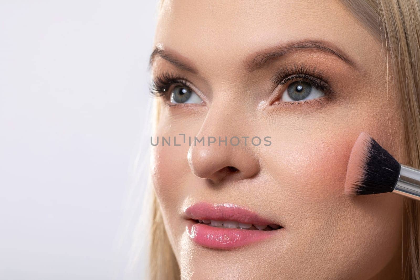 A professional makeup application showcasing a beautiful blonde woman gently applying blush with a brush