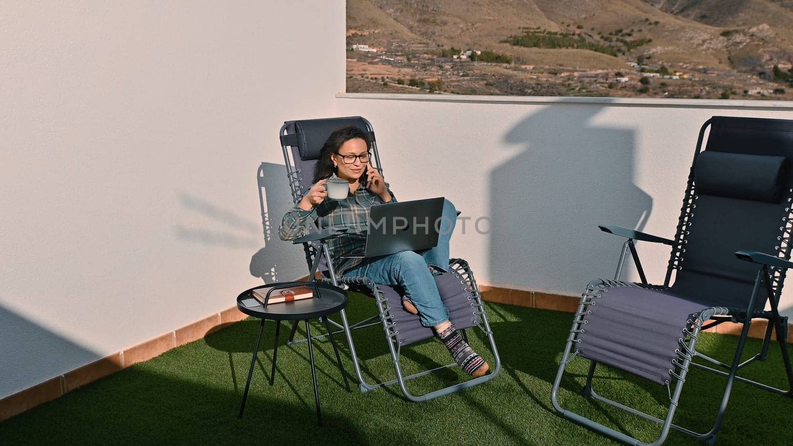 A woman enjoying remote work outdoors, sitting comfortably on a lounge chair with a laptop and coffee, basking in serene mountain views and harnessing the digital nomad lifestyle.