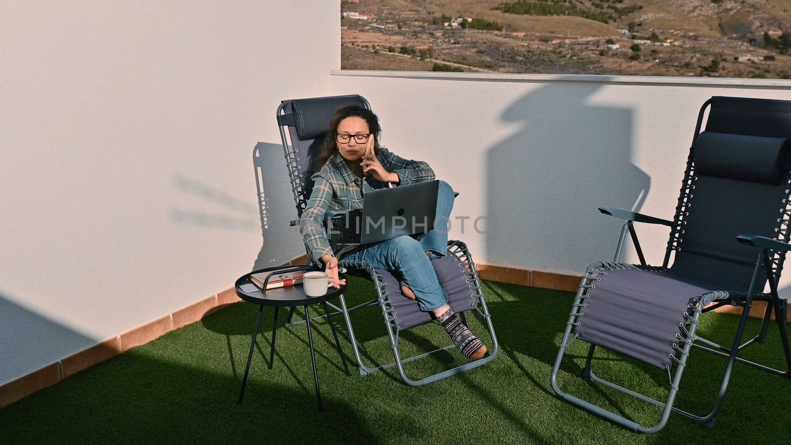 A woman working remotely on a laptop with a phone call on a sunny outdoor space.