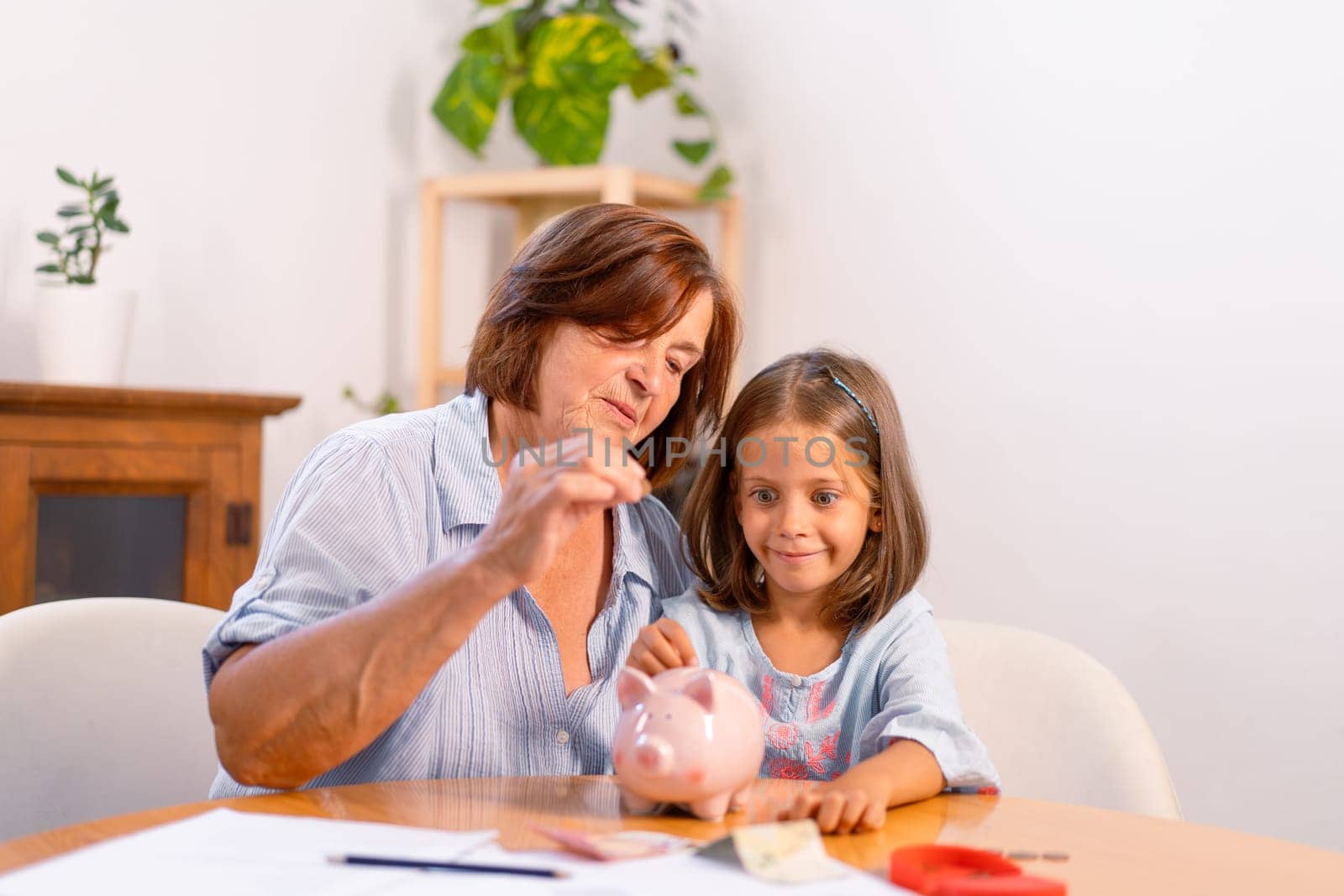 Grandmother and granddaughter inserting coins into piggy bank together on table, creating moment of bonding and teaching value of saving at home