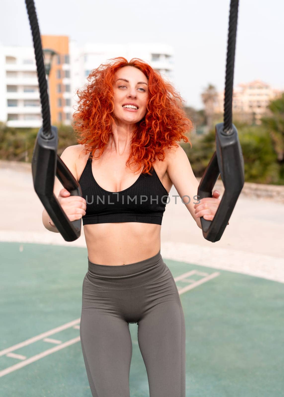 Smiling fit woman pulling elastic handles of equipment at outdoor gym. Determined young female in activewear is working out at park