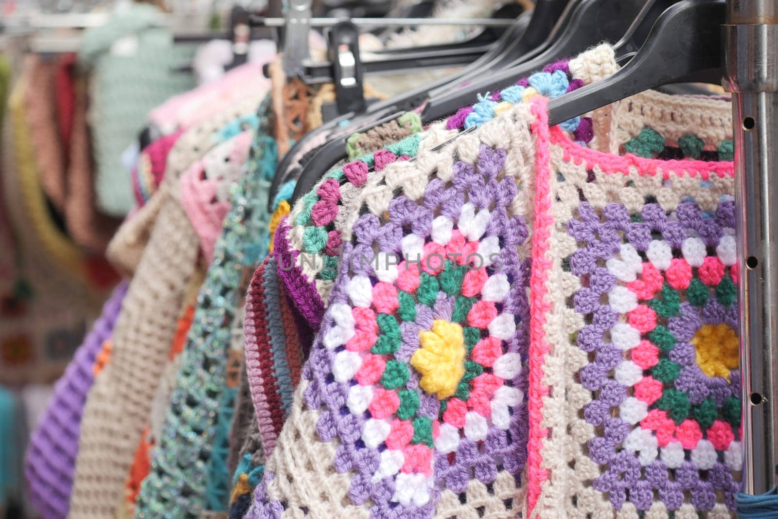Various crochet garments hang on display in a lively market setting.