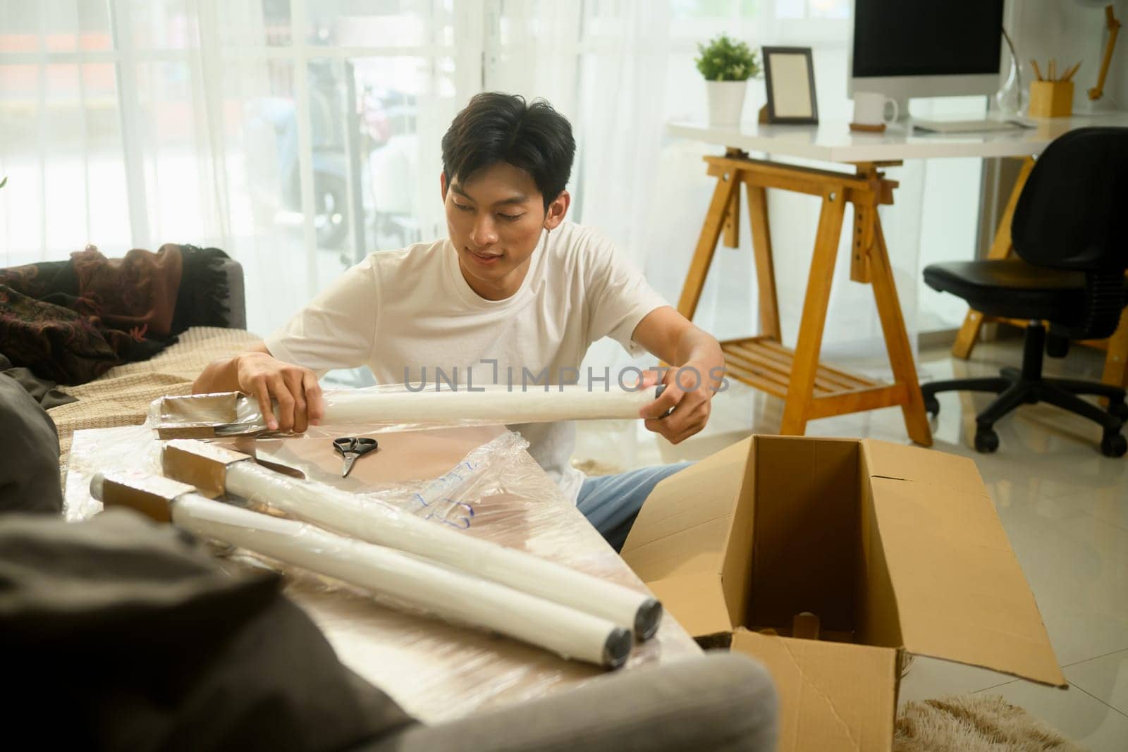 Young asian man unpacking a table leg preparing to assemble a new piece of furniture in his living room.