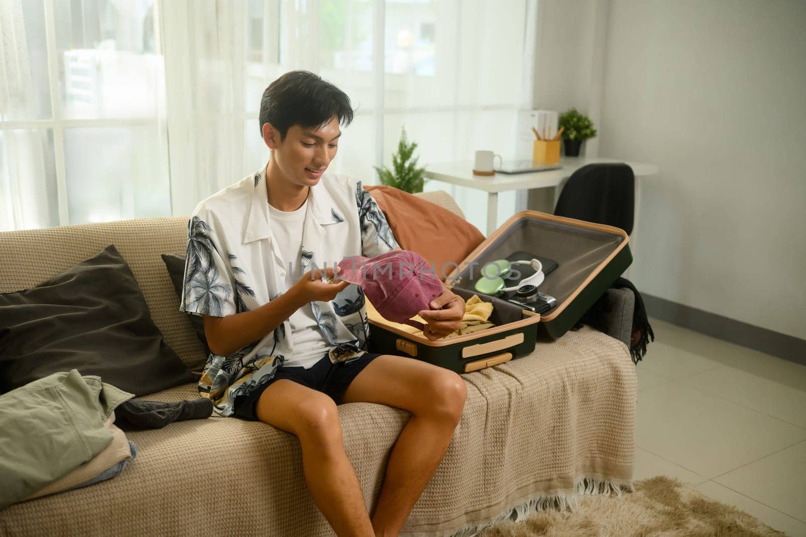 Cheerful young man in a tropical shirt packing his suitcase for summer trip.