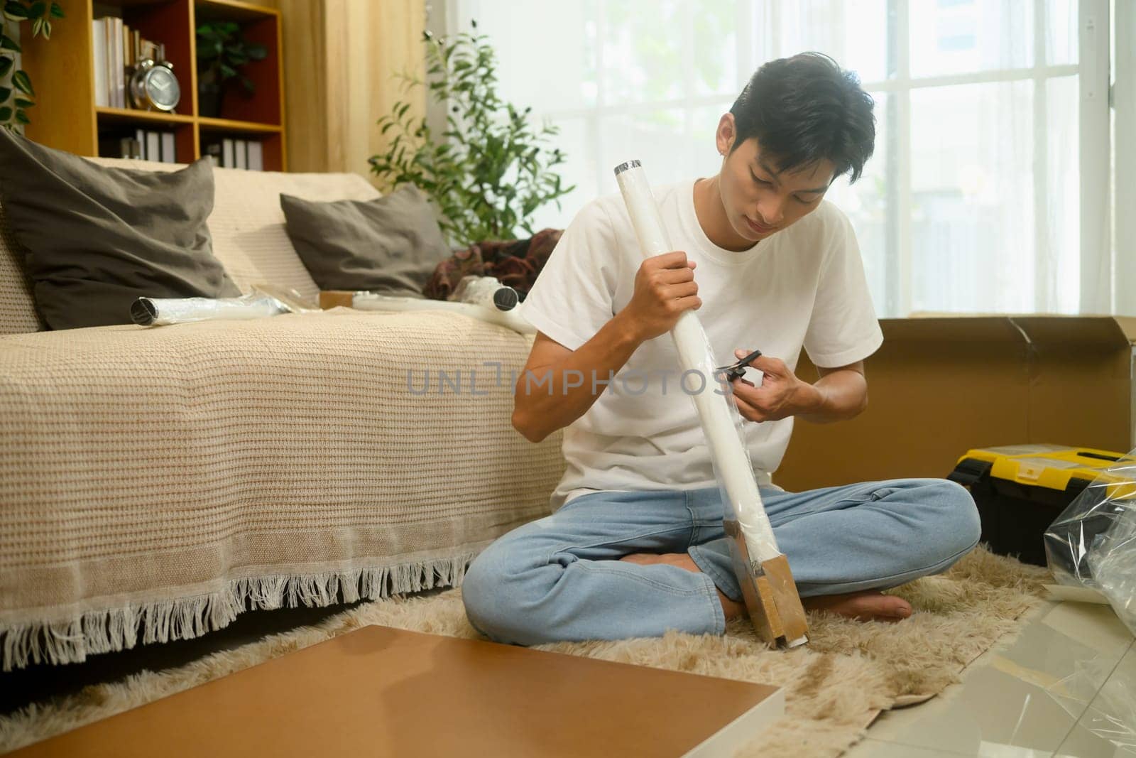 Young asian man unpacking a table leg preparing to assemble a new piece of furniture in his living room.