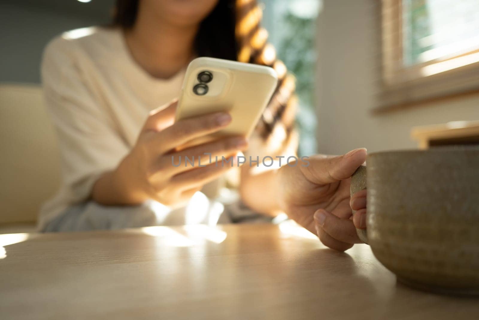 Young woman holding a cup of coffee and using mobile phone in bright living room.