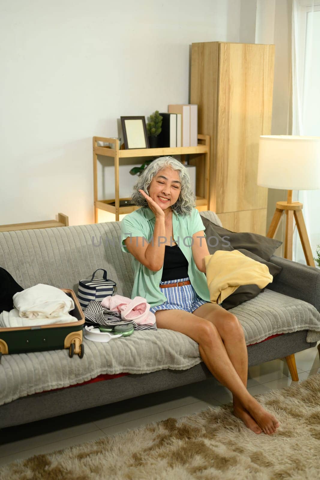 Happy senior woman is sitting on a cozy couch, preparing luggage for a summer trip.