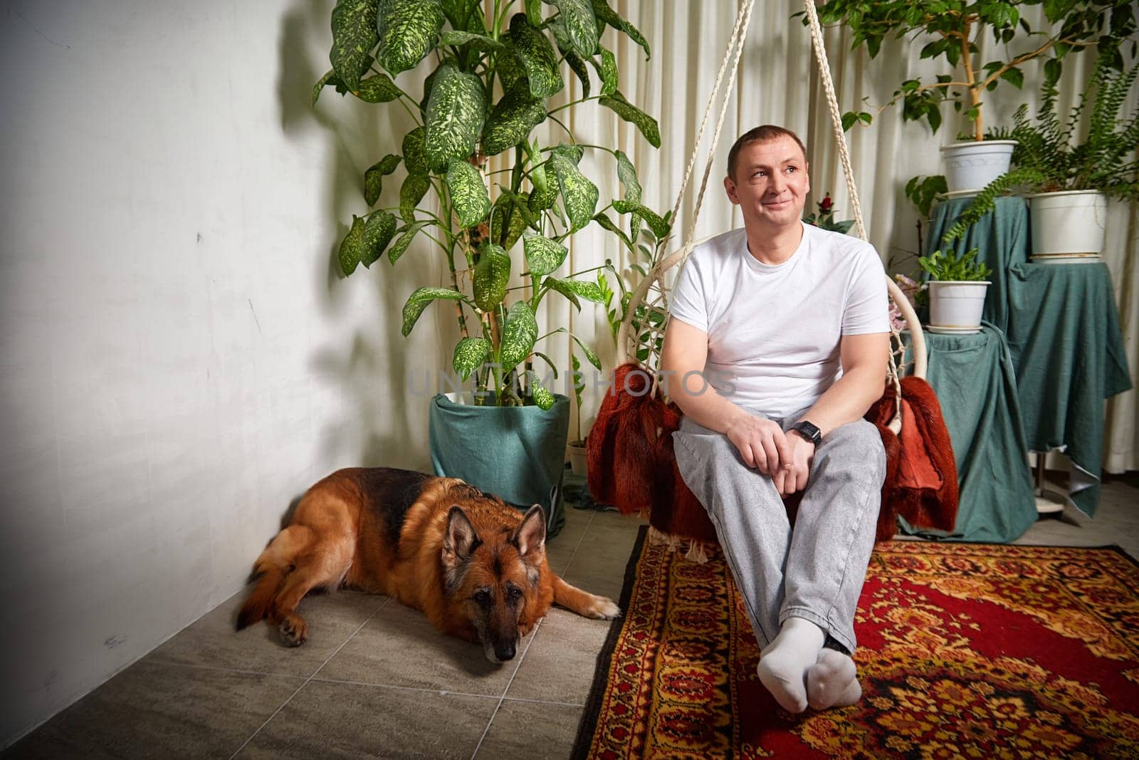 A man sits comfortably in a swinging chair while his German Shepherd lies nearby amid lush greenery.
