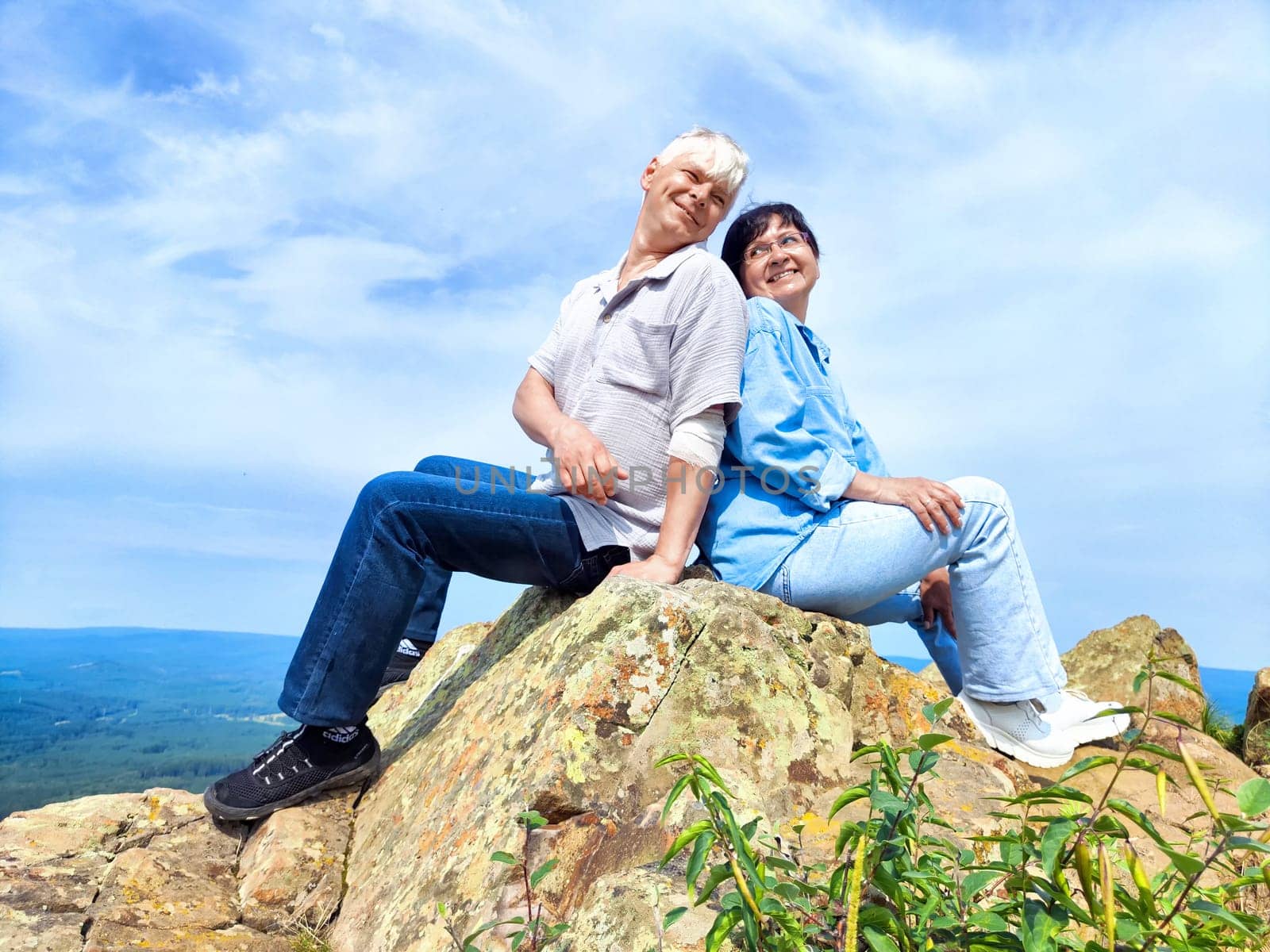 Two people sit back to back on a large rock, smiling and enjoying the beautiful view of the landscape below.