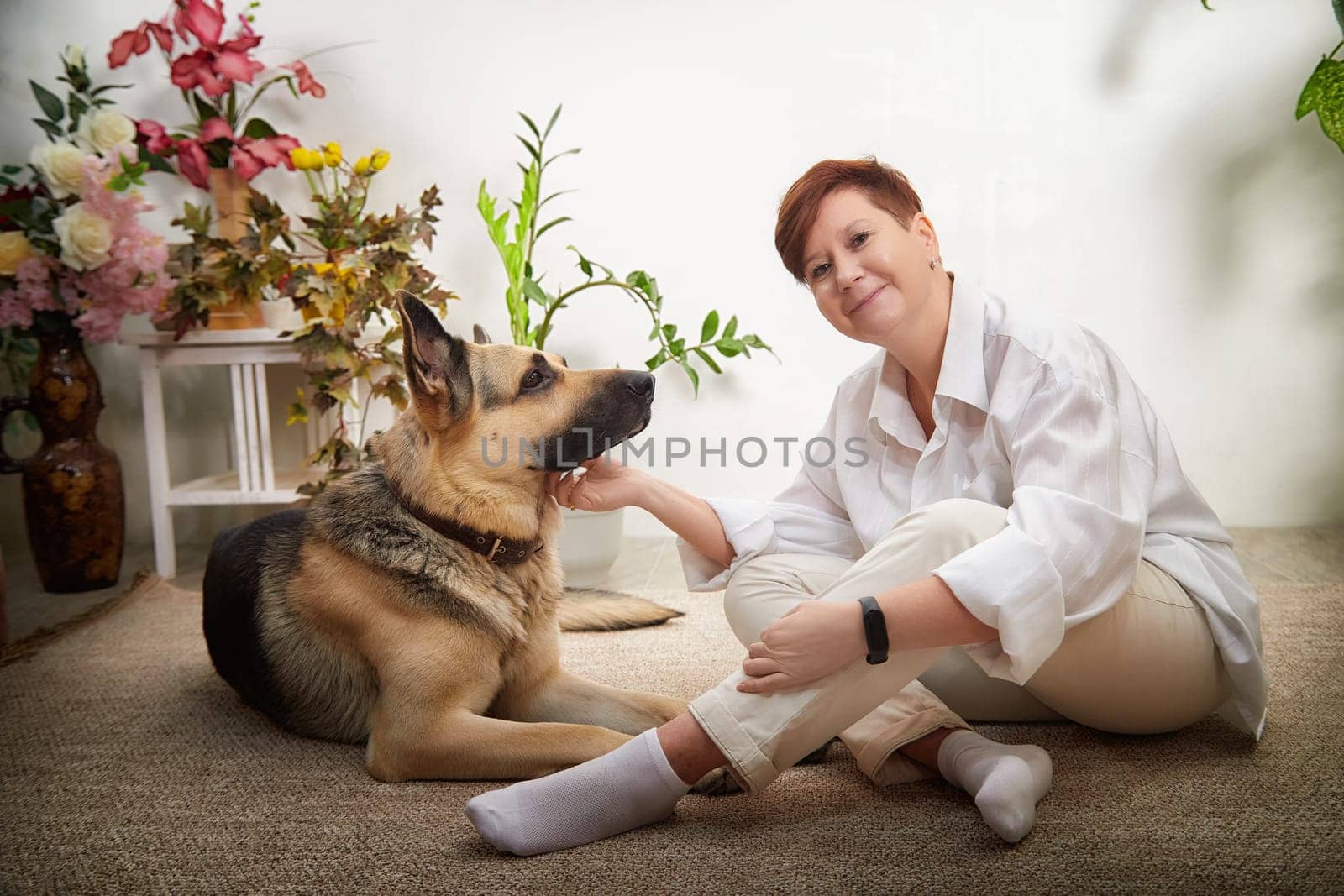 Middle-aged woman relaxes on the floor with her German Shepherd in a cozy, well-decorated room filled with plants.