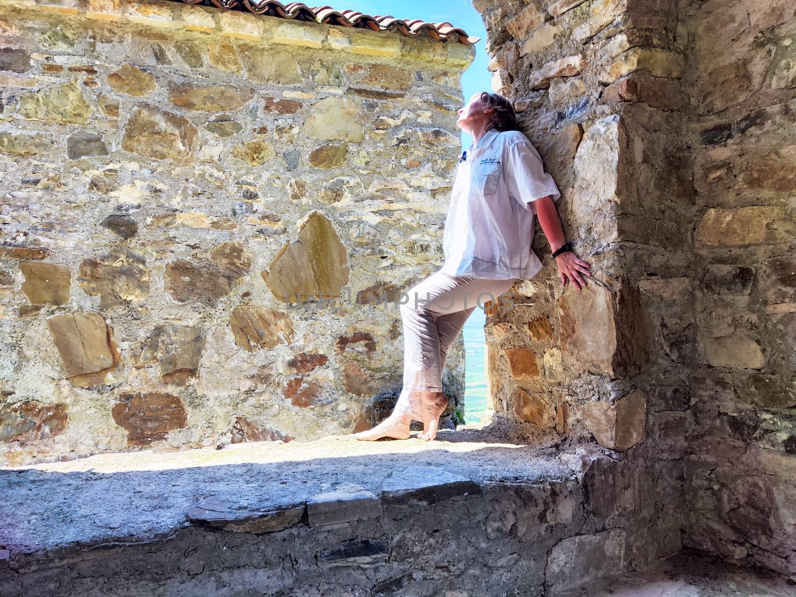A woman relaxes by a sunlit stone wall, appreciating the coastal view during a serene afternoon.
