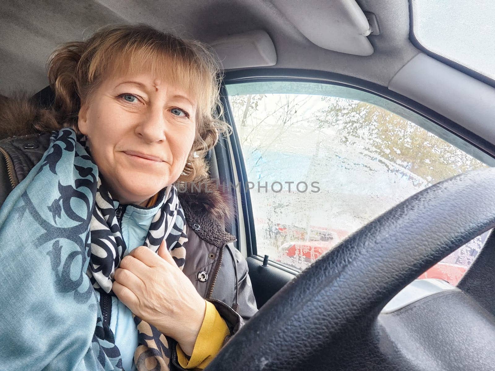 A woman smiles while seated in her car, wrapped in a warm scarf, on a chilly day in a parking area.