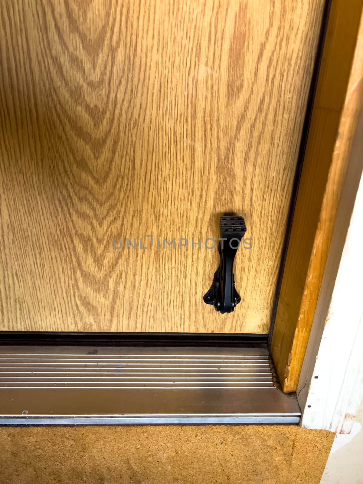 Close-up of a wooden door featuring a black metal kickstand near the bottom. The door is set within a metal threshold, and the surrounding floor has a textured surface. The kickstand is used to keep the door propped open, providing convenience and functionality.