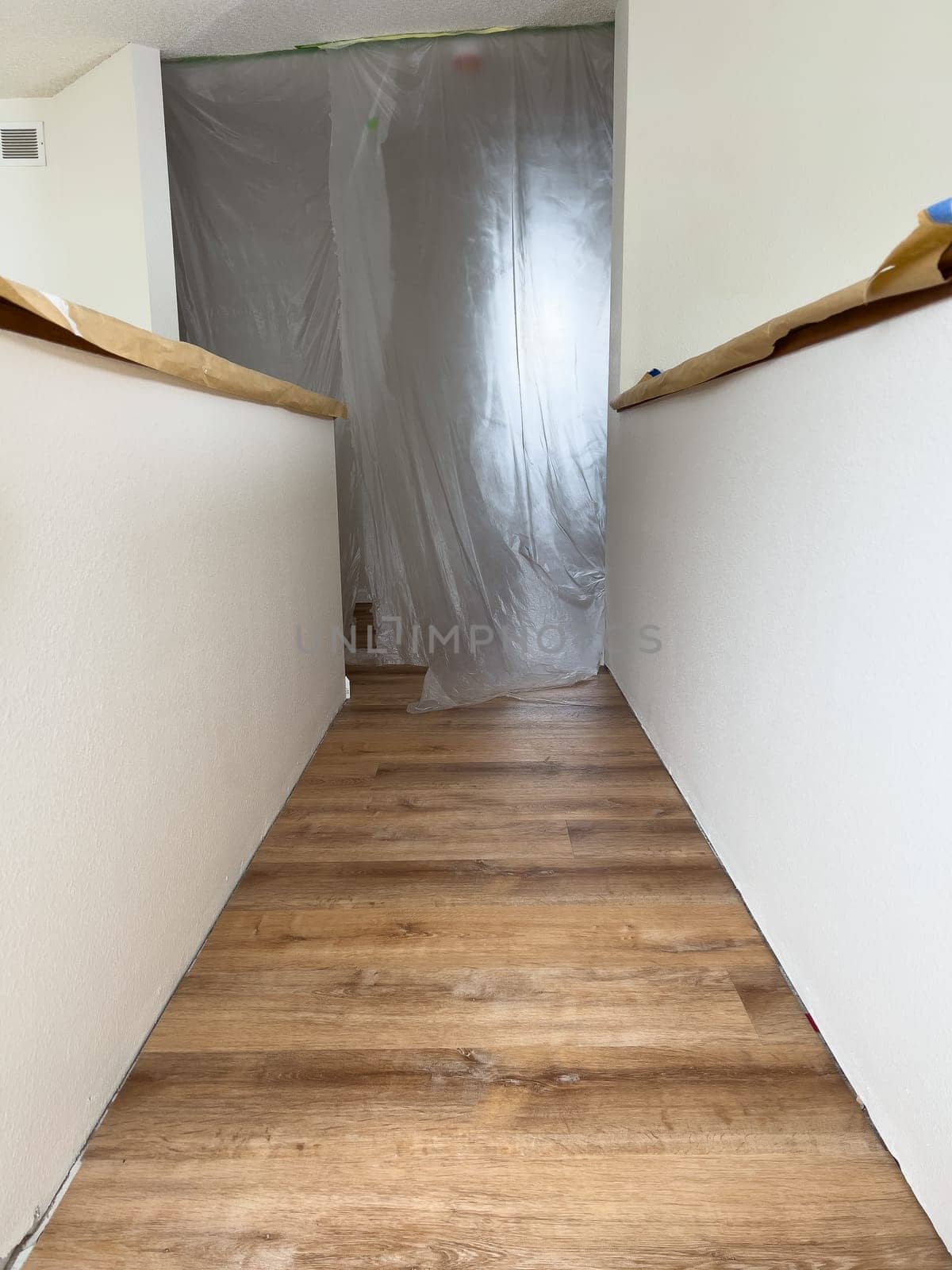 A freshly renovated upstairs hallway featuring new wood-look flooring. The walls are painted white, and protective plastic sheeting covers an entryway, indicating ongoing construction or remodeling. Handrails are partially wrapped for protection during the renovation process.