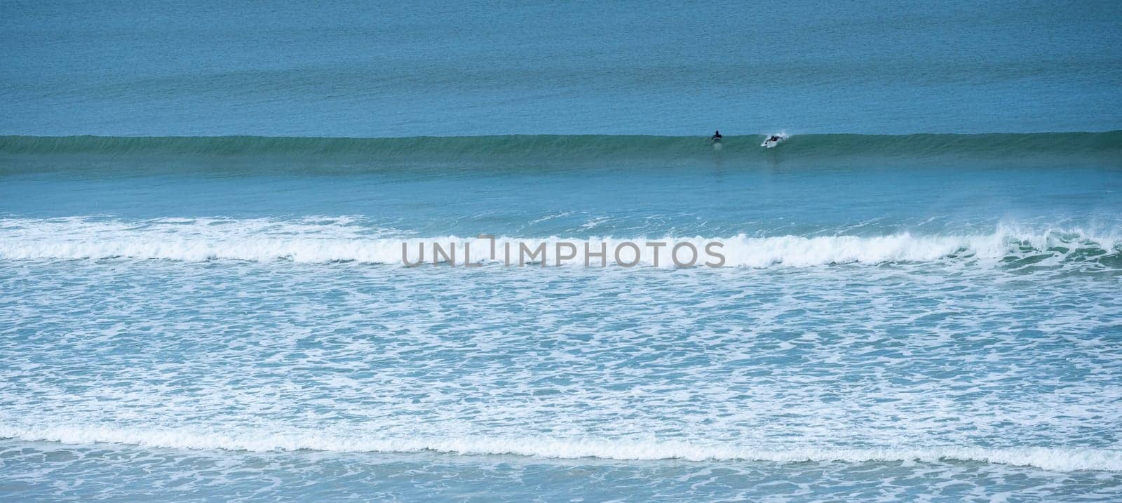 Two surfers navigate the rolling waves at Biscarrosse Beach during a serene day. The calm waters reflect the azure sky, creating a perfect surfing environment.