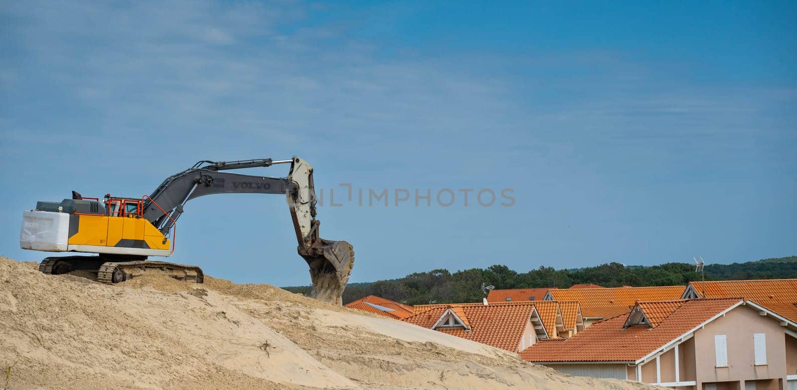An excavator moves sand along the Biscarosse coastline under a clear blue sky. Nearby houses with orange rooftops are visible, reflecting a vibrant coastal atmosphere.