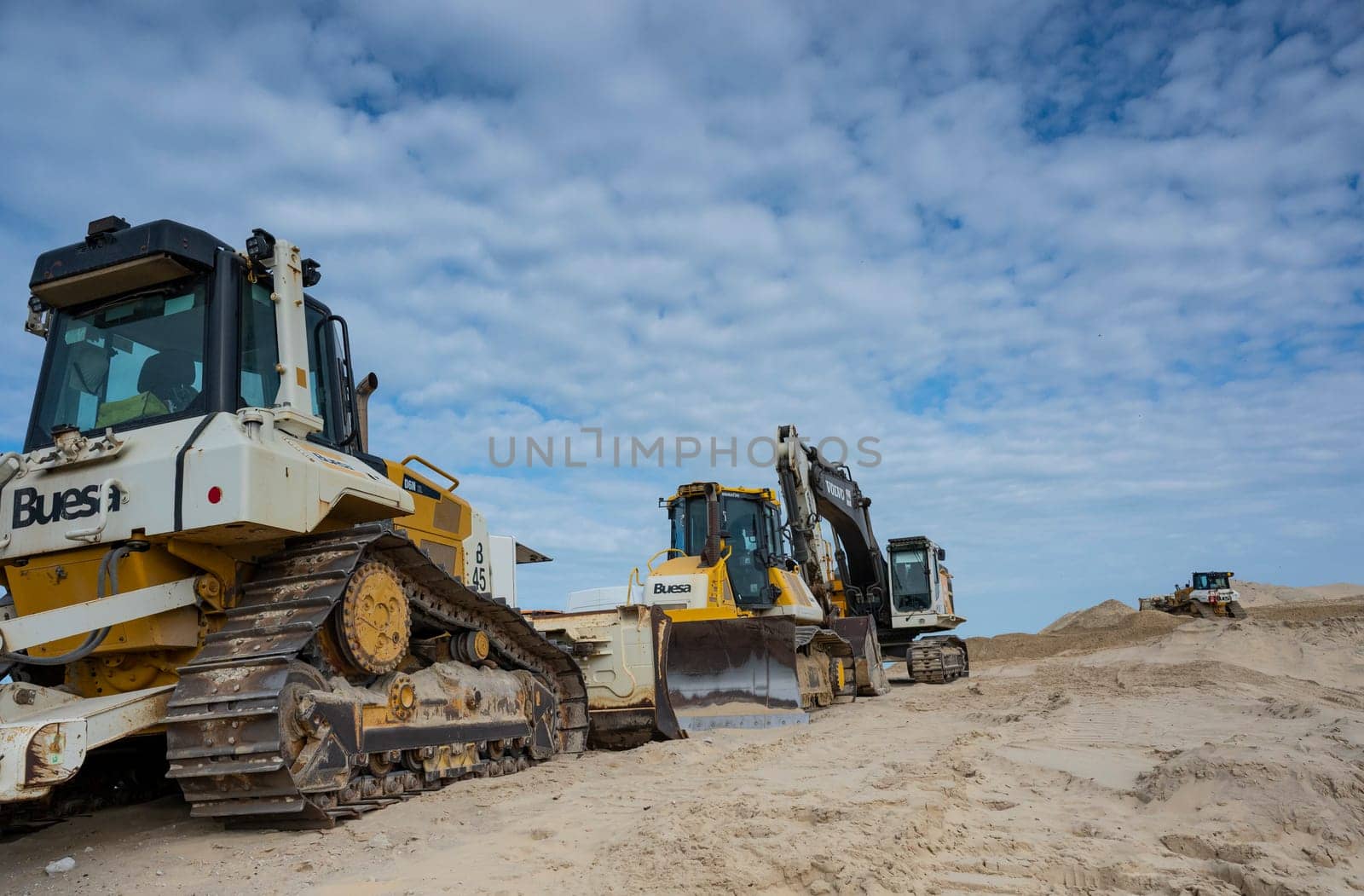 Heavy machinery is positioned along the sandy coastline, actively involved in erosion control and coastal protection efforts under a cloudy sky.