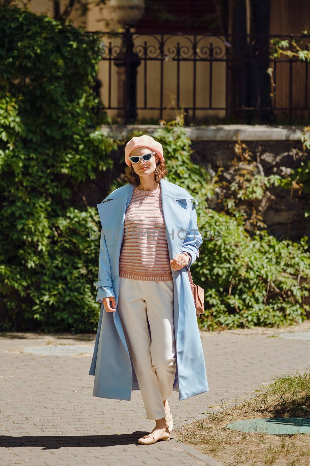 Stylish woman in blue trench coat, white trousers and coral beret walking in calm old town
