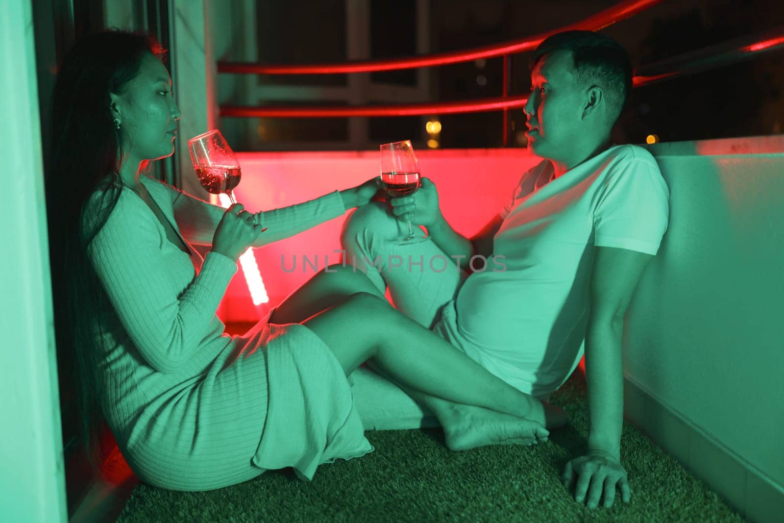 Romantic couple enjoying wine on their balcony at night, illuminated by vibrant neon lights