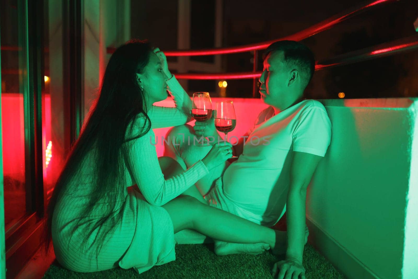 Young couple enjoying wine on their balcony at night, illuminated by vibrant neon lights