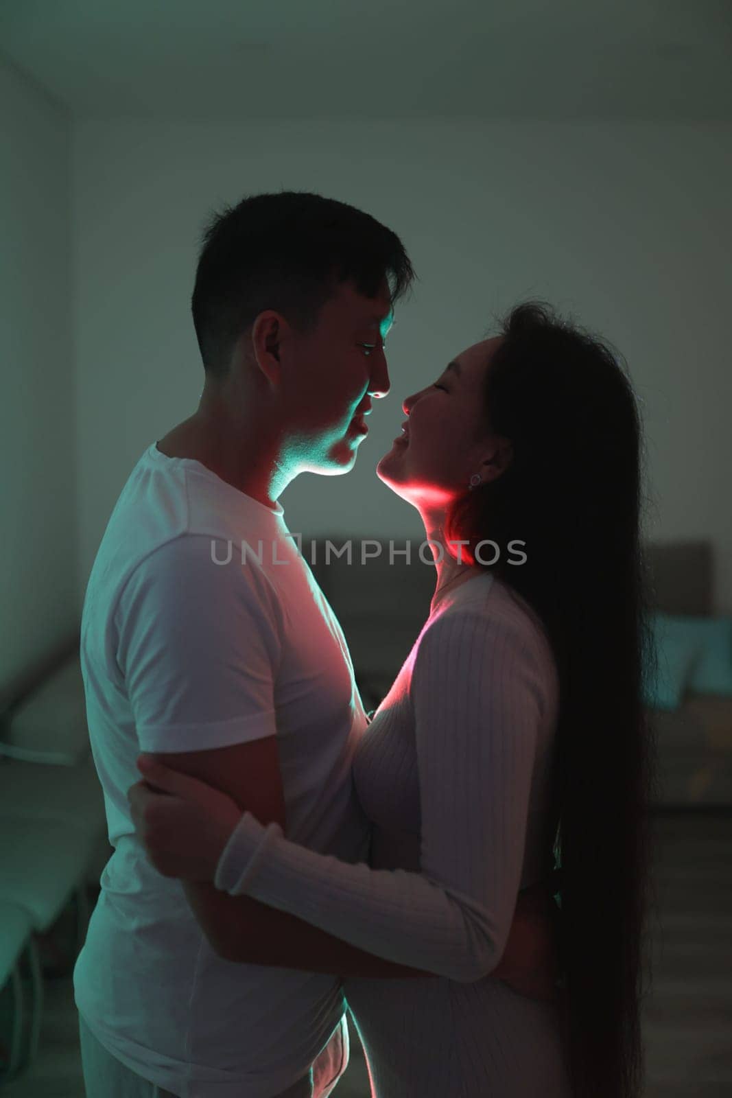 Asian couple embracing tenderly in a dimly lit room, illuminated by colorful neon lights, creating a romantic atmosphere