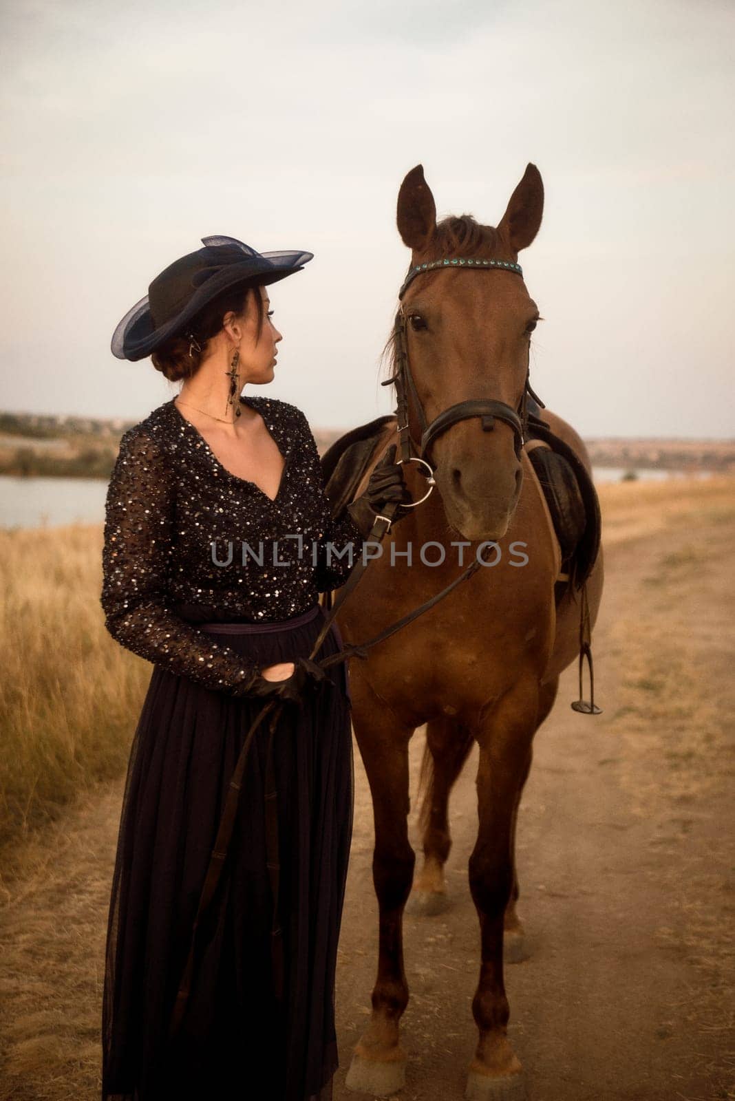 beautiful equestrian woman posing with horse after horse riding. charming girl in long dark blue dress, airy hat and gloves. leads her horse, by the reins, in the field in the rays of the setting sun. concept of women riding horses