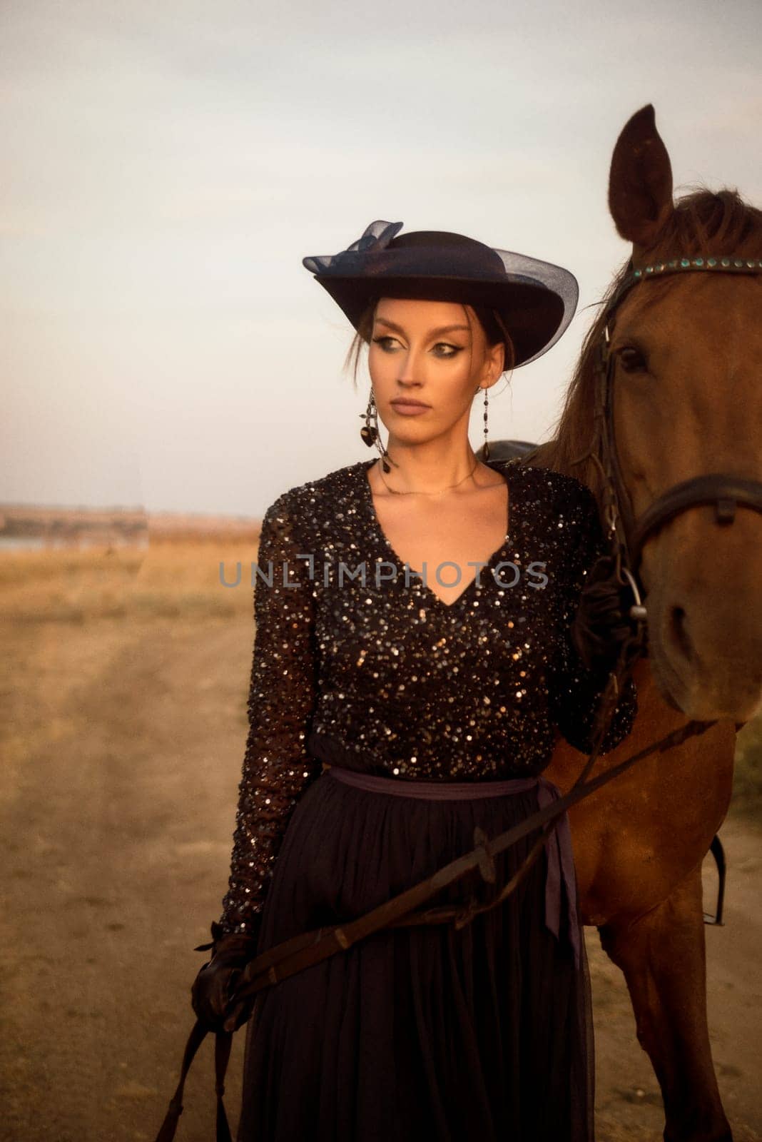close-up of beautiful equestrian is resting after horse riding. charming girl in long dark blue dress, airy hat and gloves. leads her horse, by the reins, in the field in the rays of the setting sun. concept of women riding horses