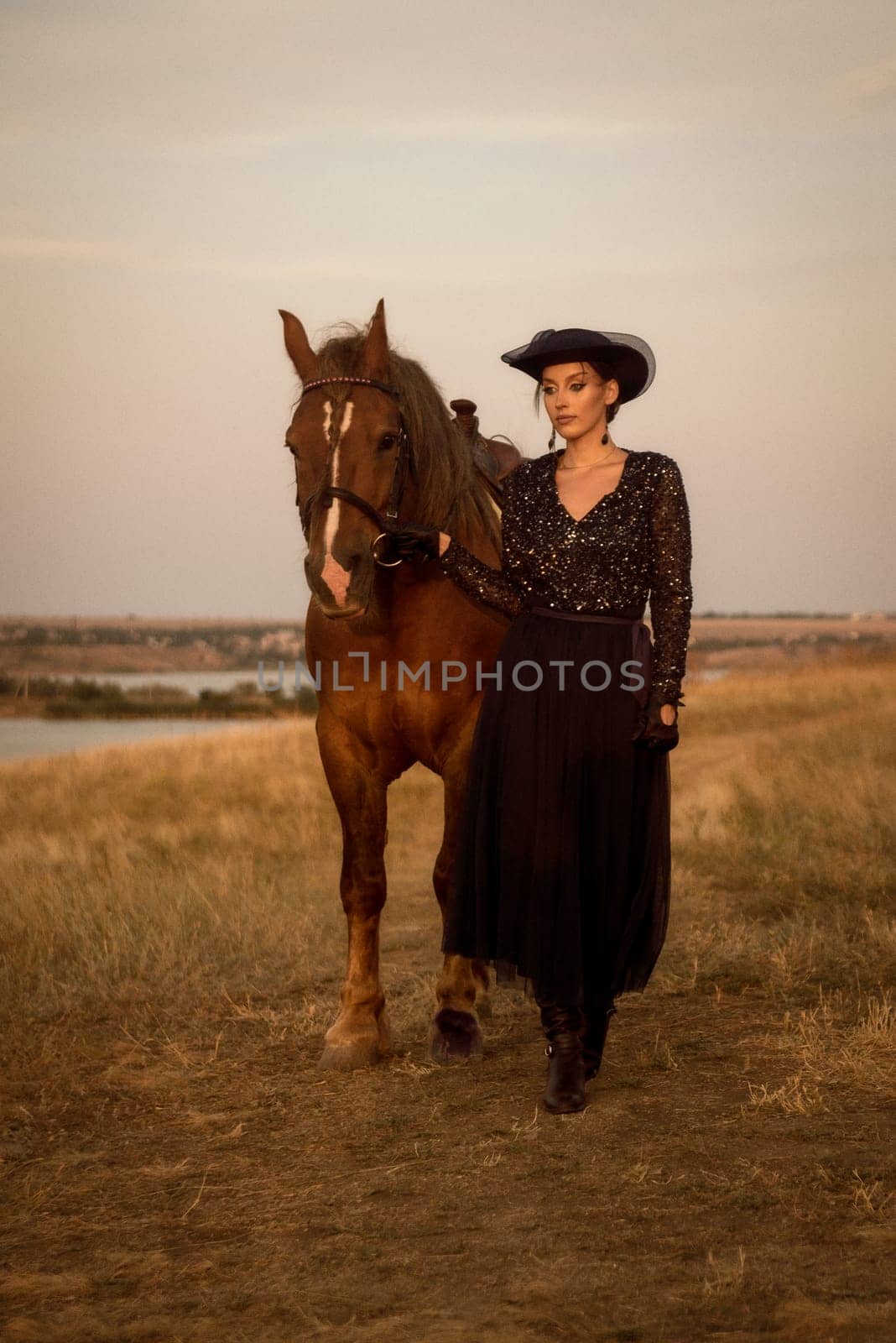 beautiful equestrian is resting after horse riding. charming girl in long dark blue dress, airy hat and gloves. leads her horse in the field in the rays of the setting sun. concept of women riding horses