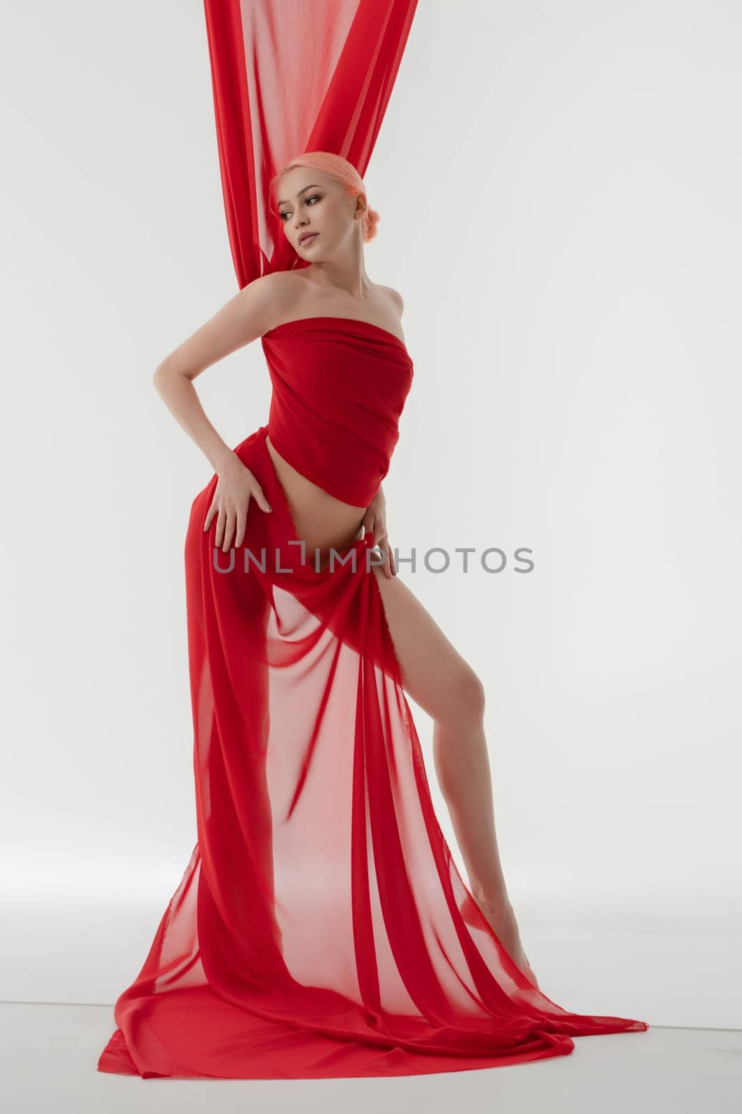 A nude woman wrapped in light red fabric poses gracefully, showcasing her beauty and elegance in a minimalist setting.