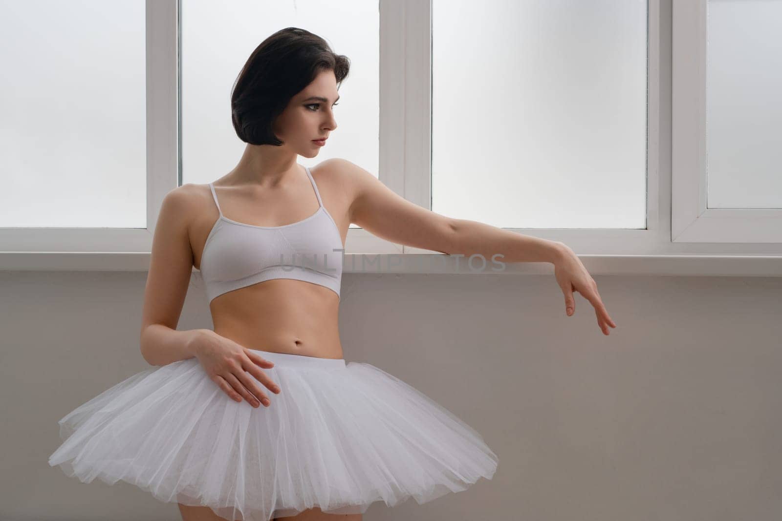 A woman wearing lingerie and a tutu demonstrates ballet poses in a bright studio.