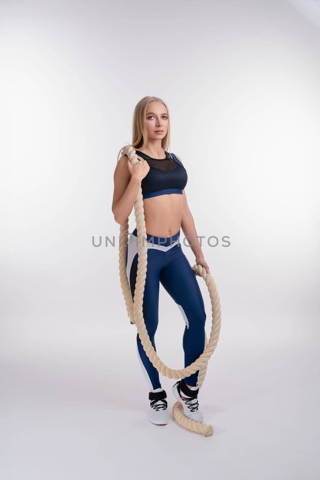 A woman stands confidently in a studio, holding battle ropes, showcasing her strength and beauty in athletic wear.
