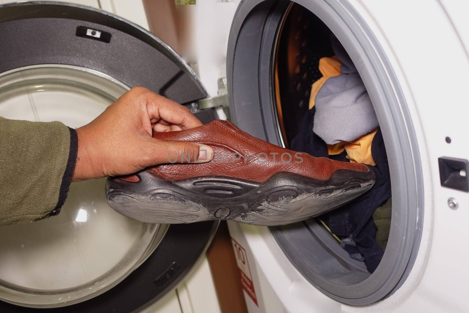 A person inserts a brown shoe into a washing machine filled with clothes.