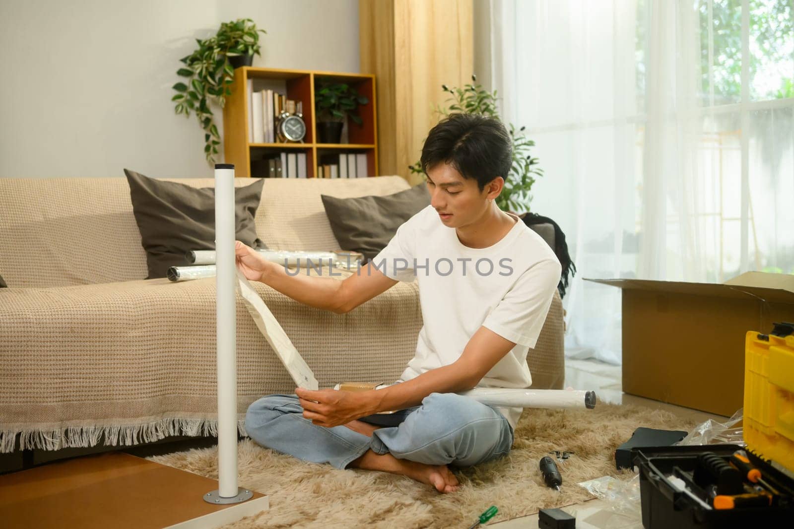 Young man reading an instruction manual while assembling furniture in a bright living room.