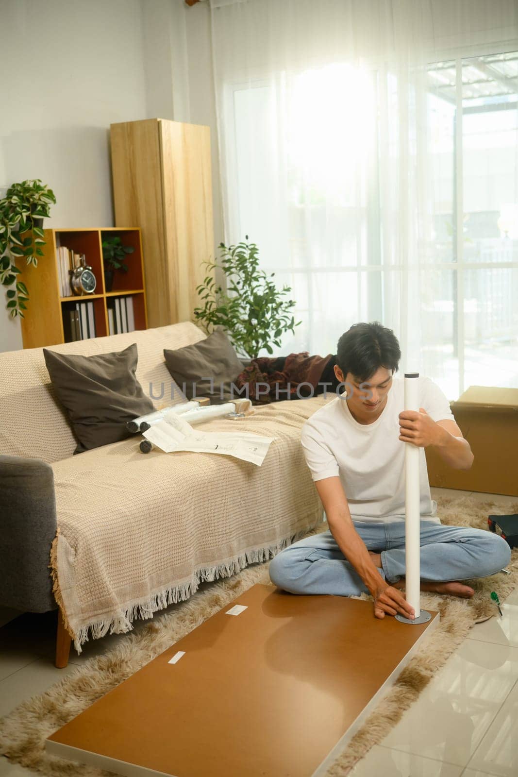 Young asian man sitting on soft rug assembling a table and following assembly instructions.