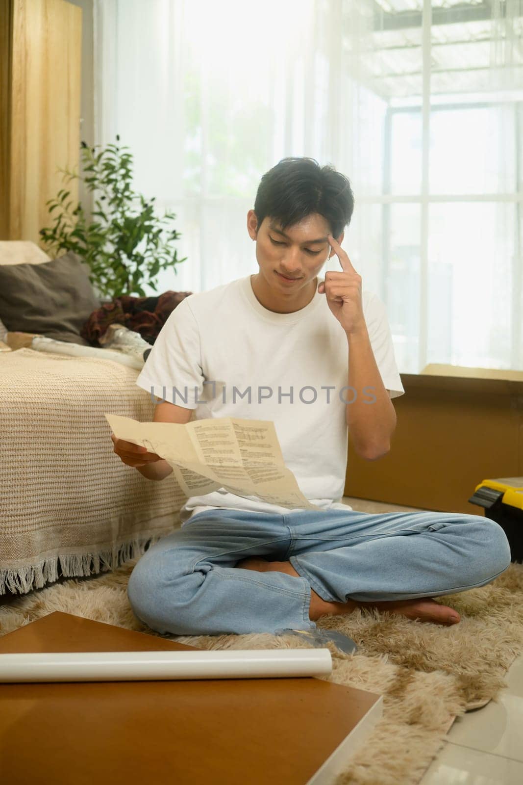 Young man reading an instruction manual while assembling furniture in a bright living room.