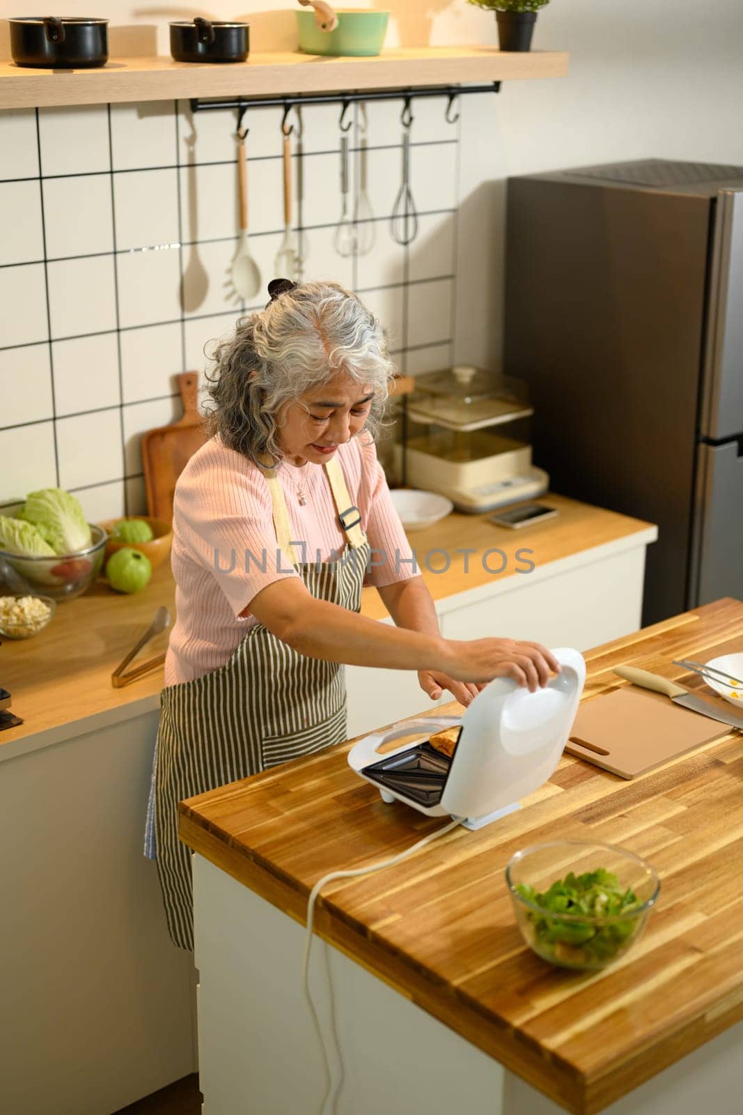 Happy senior woman making a delicious toasted sandwich for breakfast in a kitchen.