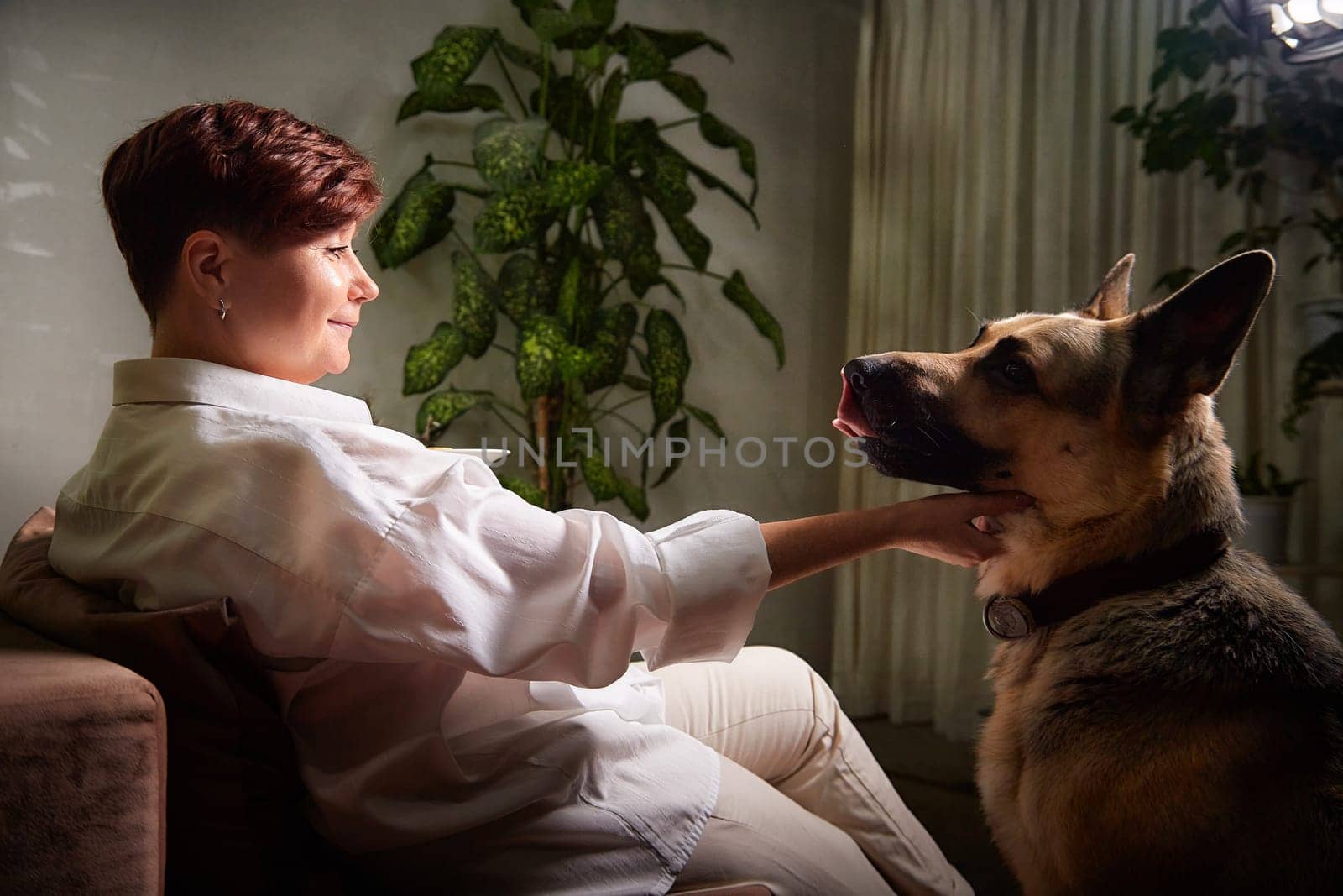 Blonde woman interacts with her German Shepherd in a warm, inviting space filled with plants.
