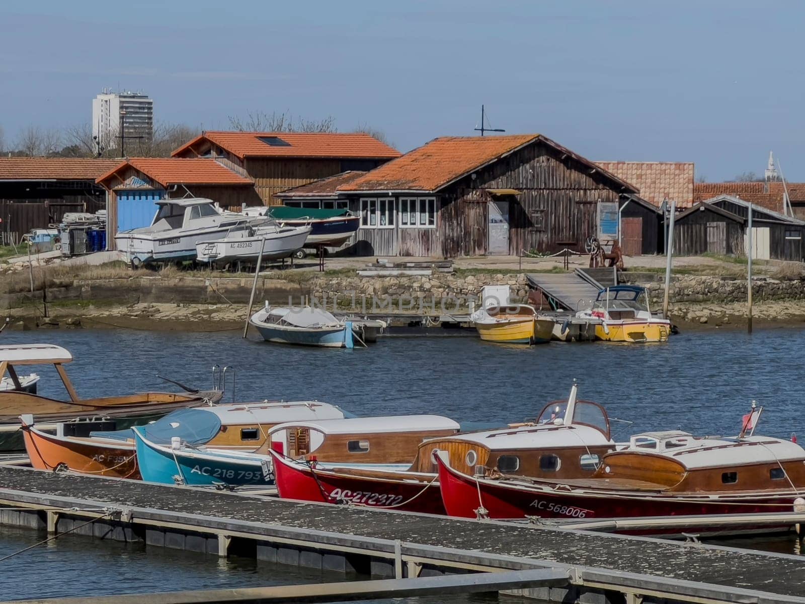 Boats line the dock in Port La Teste de Buch, showcasing various colors under a clear blue sky, while rustic buildings provide a charming backdrop to the tranquil waterfront setting.