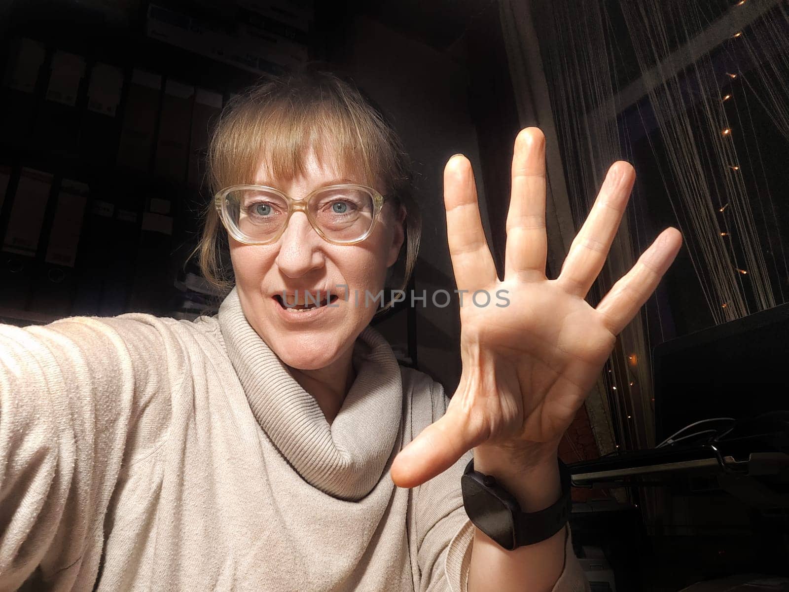 A middle-aged woman takes a fun selfie, showcasing her playful personality and cozy indoor environment at dusk.