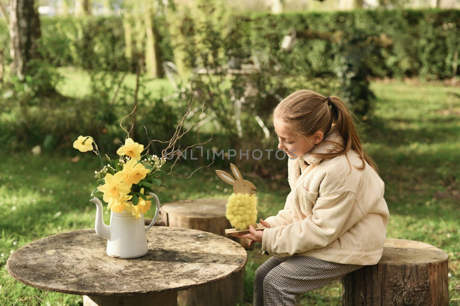 A girl in a garden holds a yellow woolen rabbit in her hands. On a wooden table is a vase with daffodils. Easter