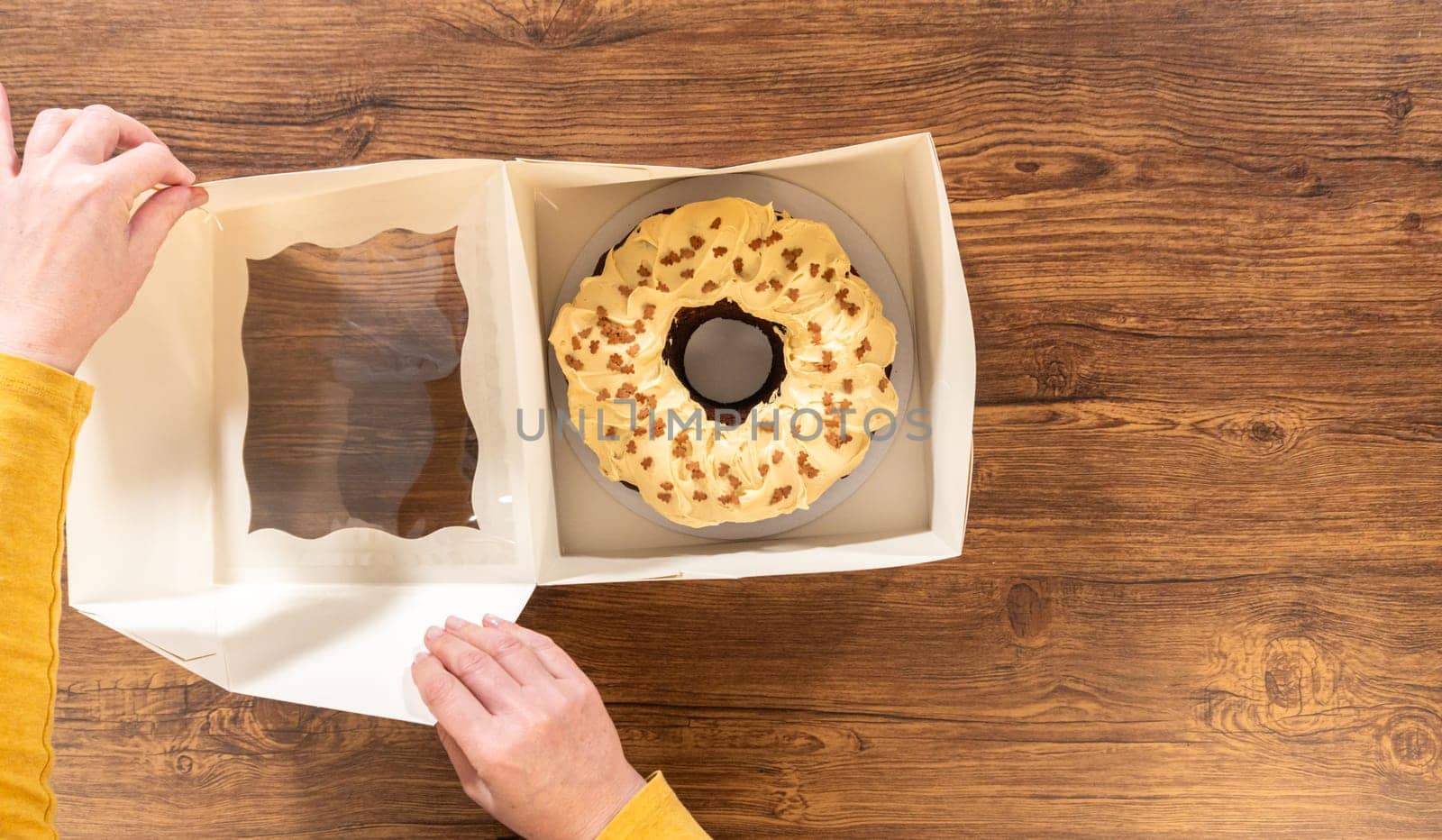 Flat lay. Carefully placing the gingerbread bundt cake, adorned with salted caramel frosting and gingerbread sprinkles, into a white paper bundt cake box with a clear window for gifting.