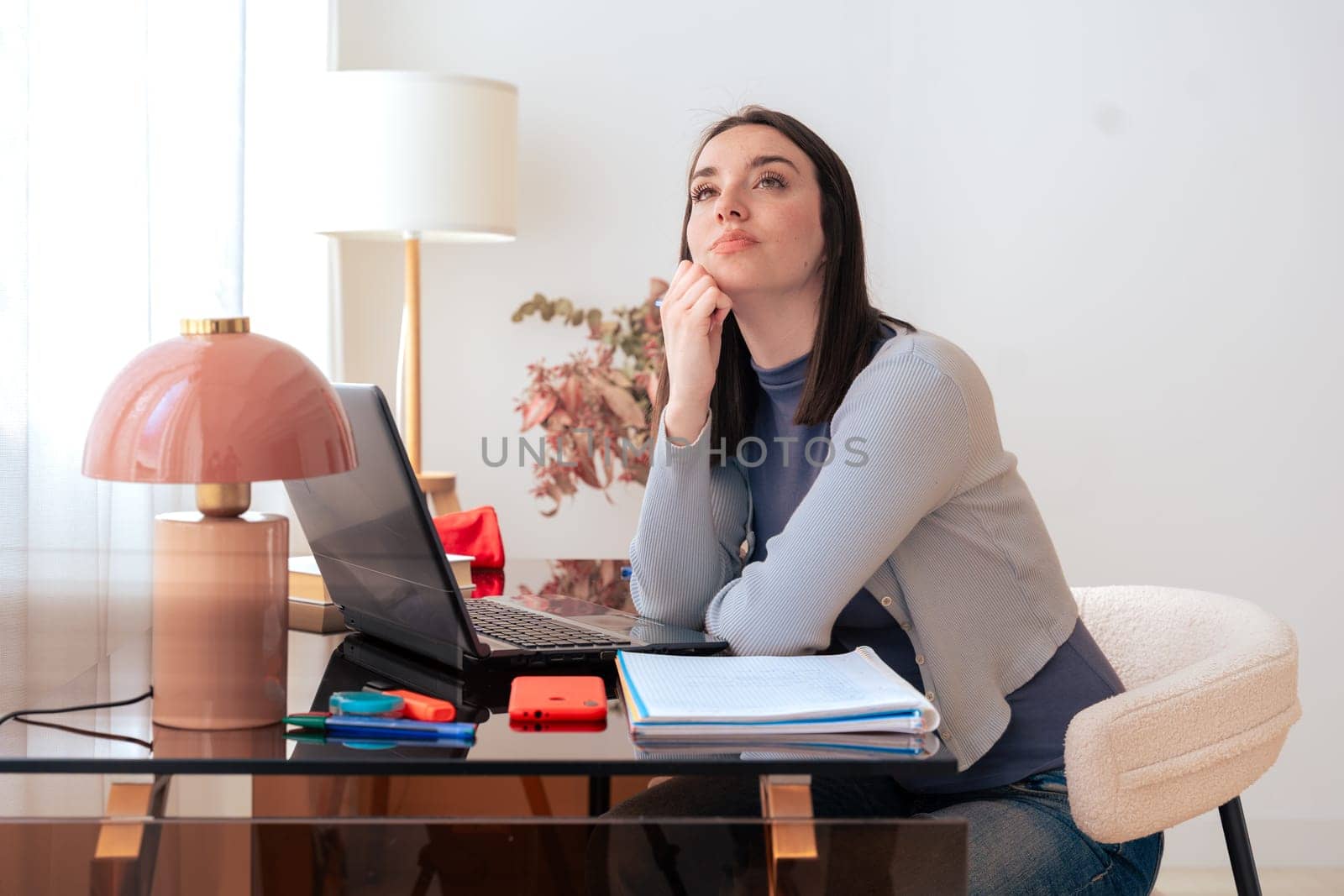 Caucasian woman sitting at desk, thinking while looking up. Laptop, notebook, and stationery on desk in modern home office. Creativity, concentration, decision-making, productivity, and contemplation.