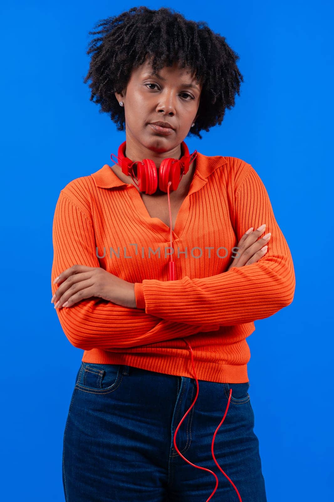 Serious African American woman wearing red headphones, posing confidently with arms crossed against a blue background. Modern fashion, casual style, technology, self-assurance, and positivity.