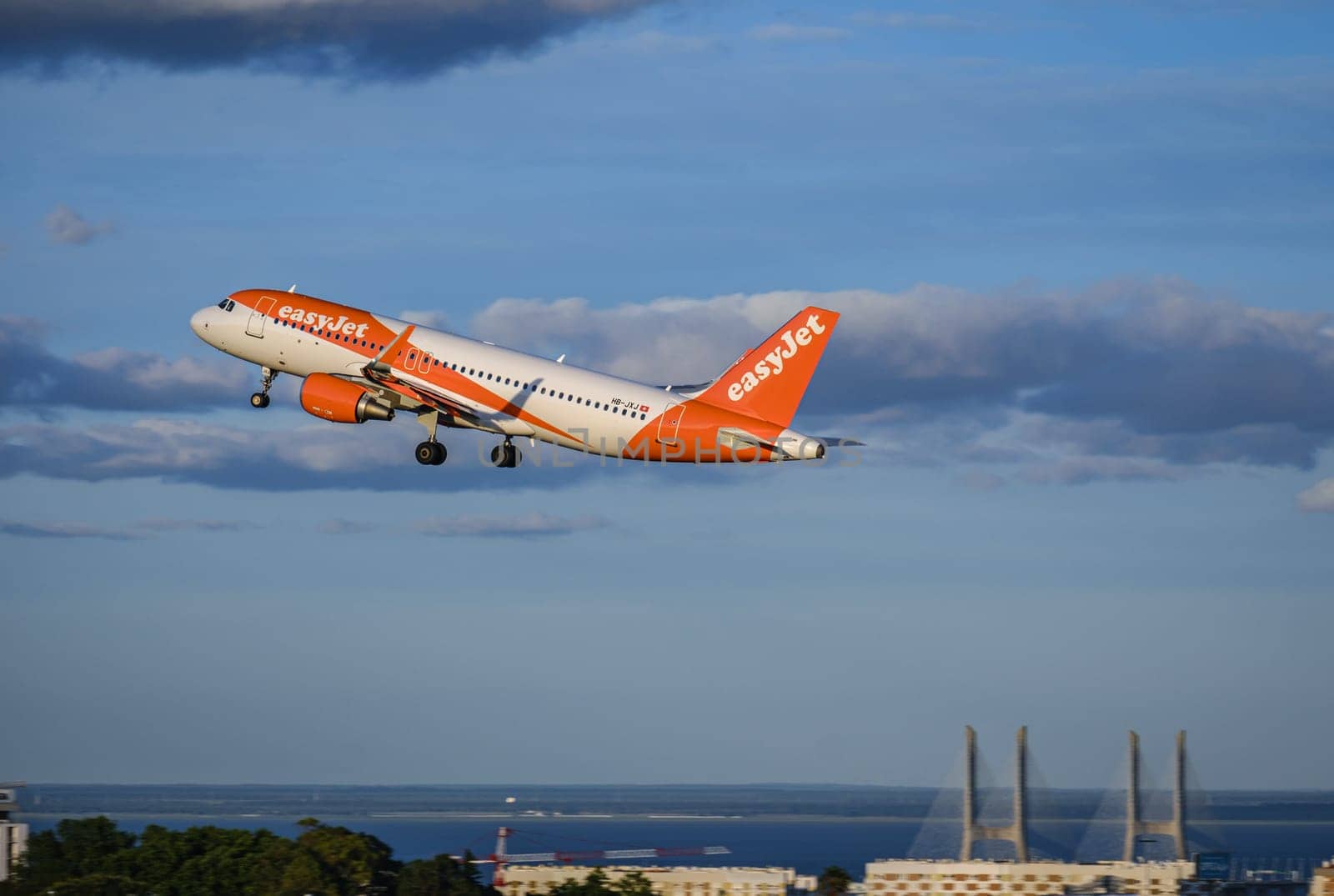Lisbon, Portugal - Apr 29, 2024: Panning shot of EasyJet Airbus 320 aircraft take off from Lisbon airport runway. Cityscape, river view in background, airport warehouse facility, low-cost airline in clear blue sky and some clouds on sunny day