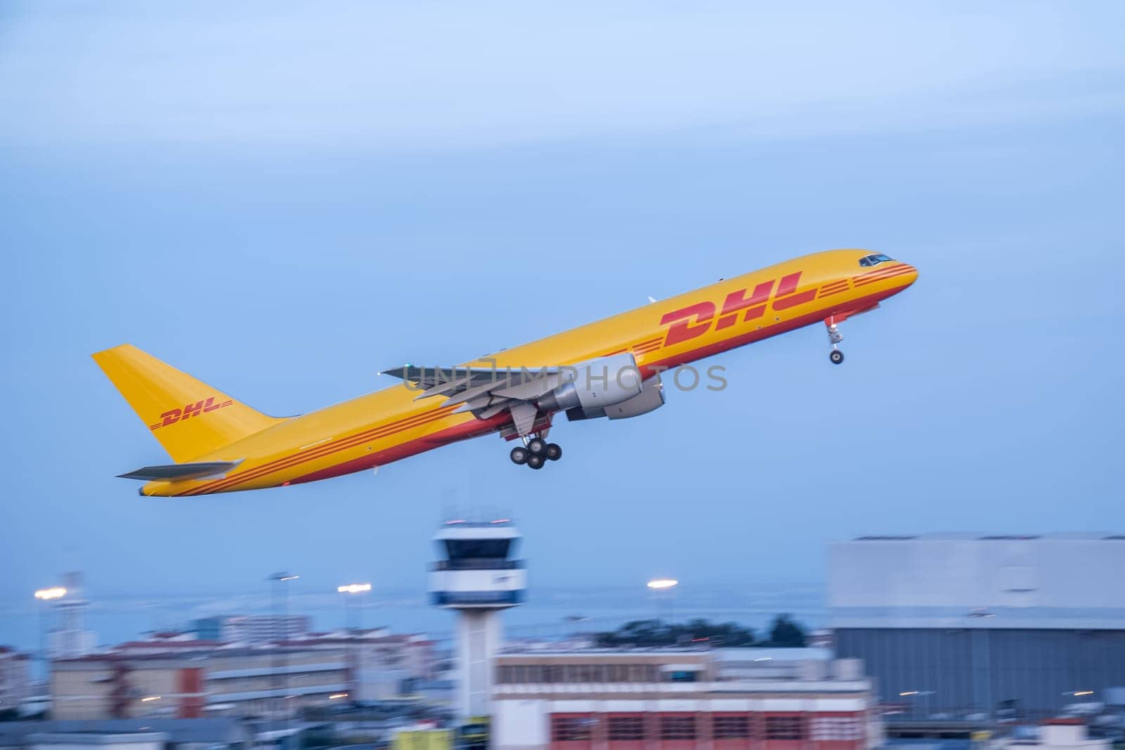 Lisbon, Portugal - Apr 3, 2024: Panning shot of DHL cargo airplane Boeing 757-200F during takeoff from airport, with city buildings and control tower in background at dusk. Blurred background, air freight, delivery service, international logistics,